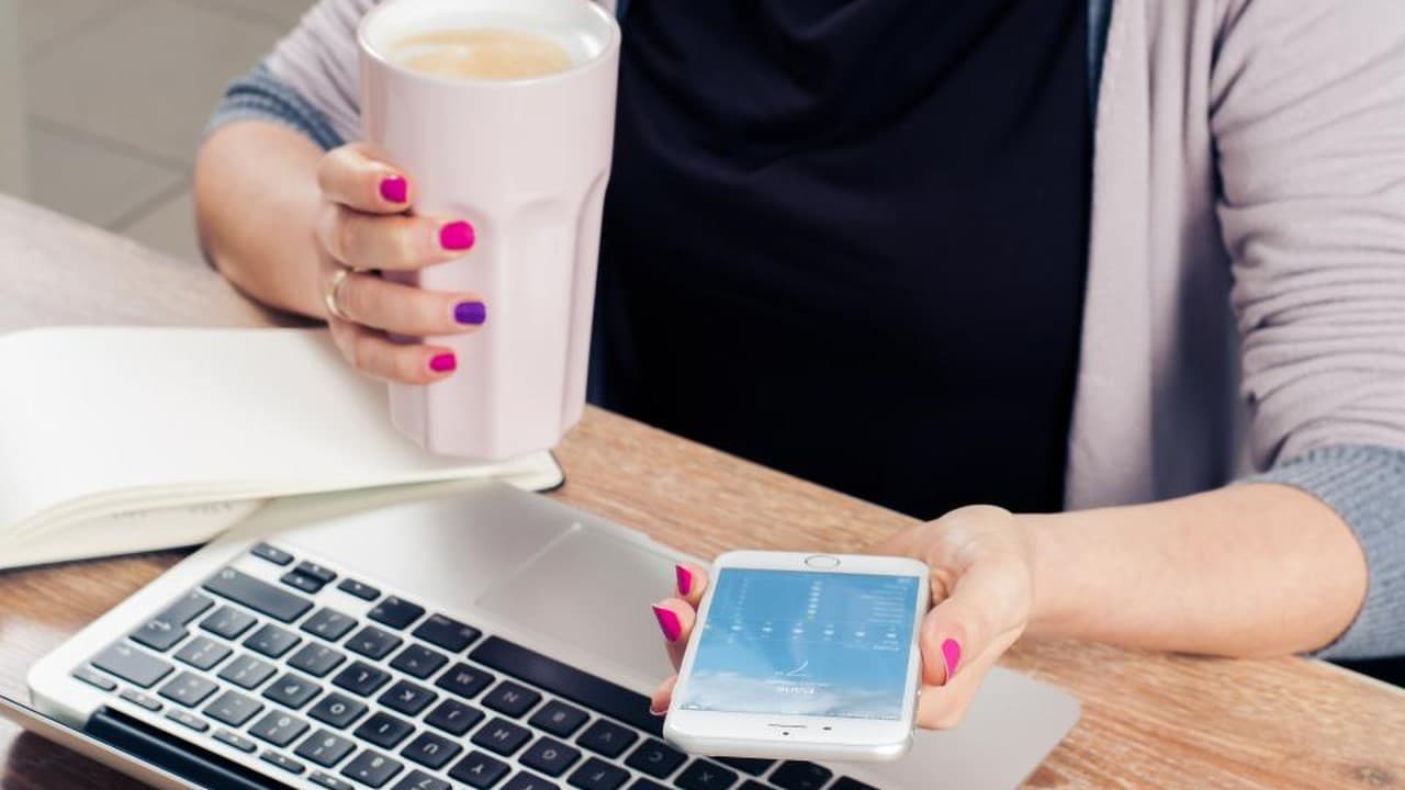 Woman holding a phone and coffee beside a laptop and notebook