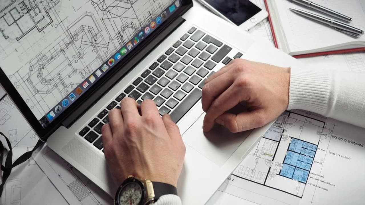 Hands typing on a laptop next to printed design plans